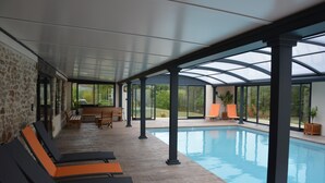 Un espace piscine couvert comprenant des chaises longues, une table en bois et une vue sur l’extérieur.