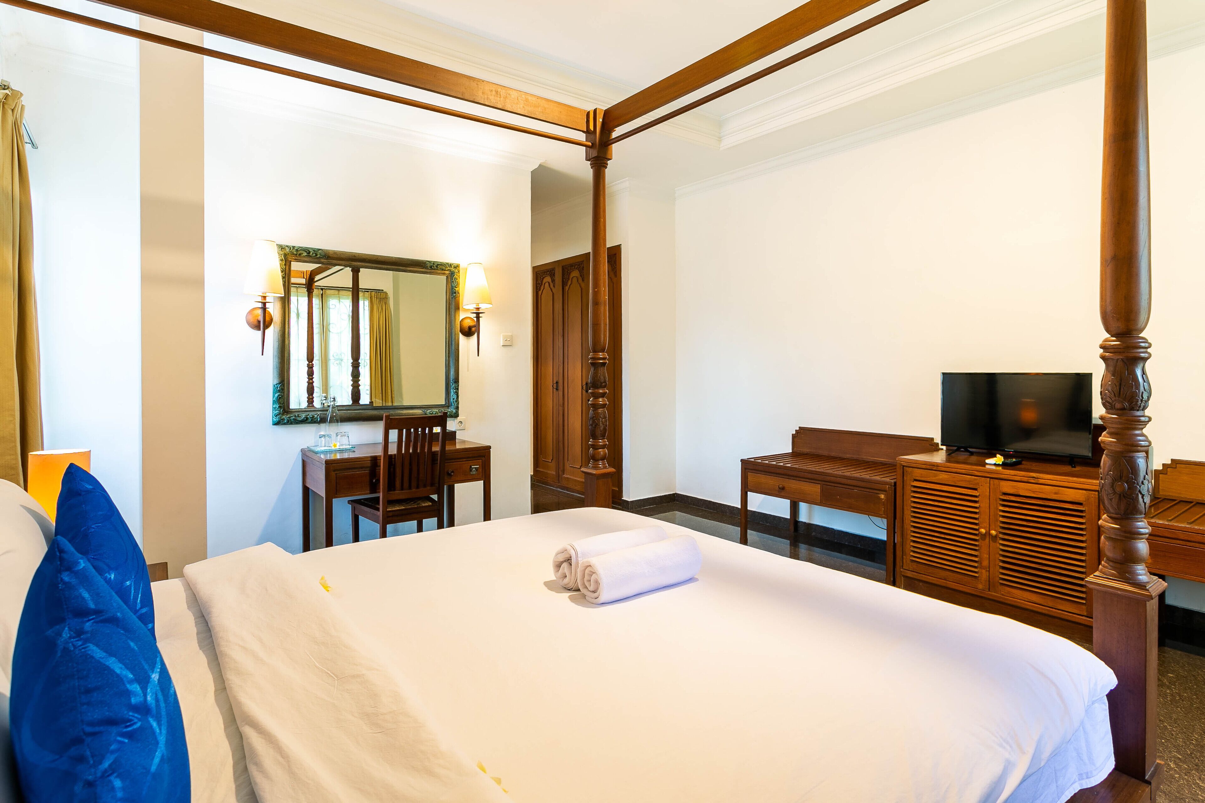 A hotel room with a four-poster bed, a desk with a chair, a television, and a mirror.