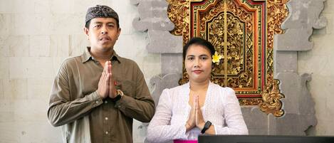 Un hombre y una mujer en posición de oración frente a un escritorio con una computadora portátil, frente a una pared decorativa con un marco dorado.