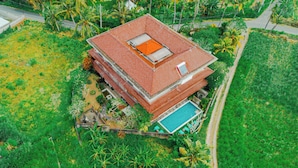 An aerial view of a multi-story building with a red tiled roof, surrounded by lush greenery and palm trees.