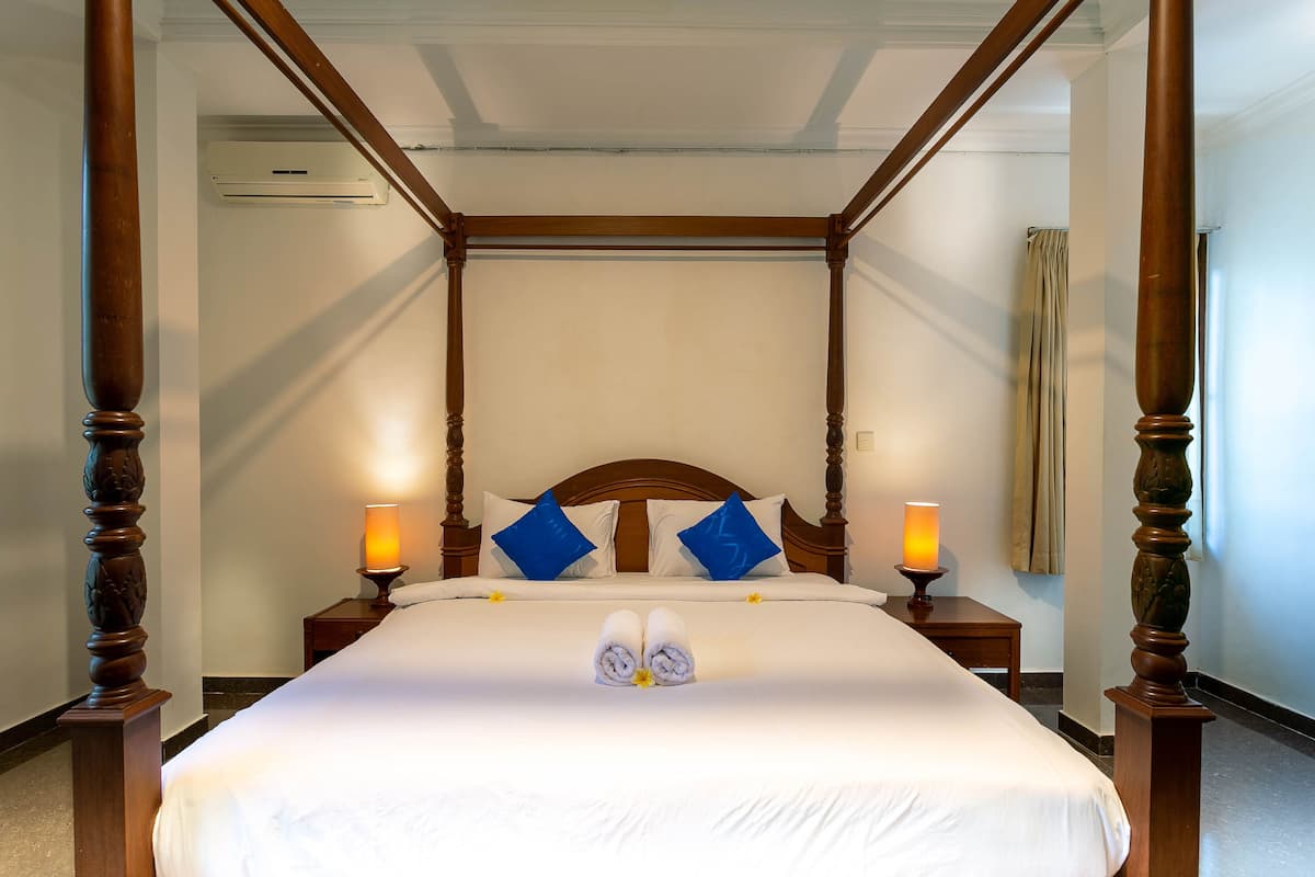 Una cama con dosel, cabecera de madera, dos mesitas de noche con lámparas y un par de toallas dispuestas sobre la cama.