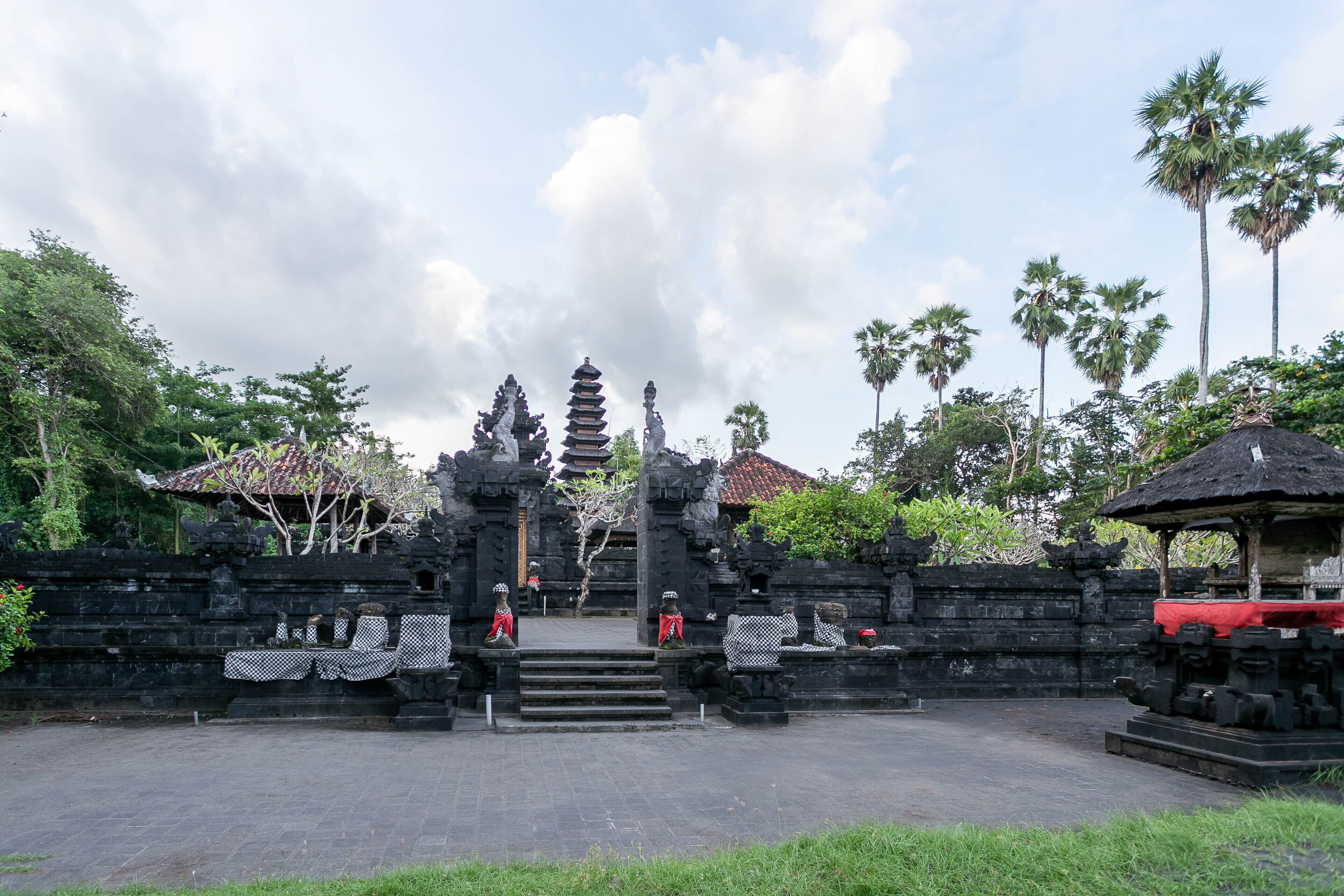 A traditional Balinese temple with a central staircase leading to a tiered roof structure, surrounded by stone walls and palm trees.