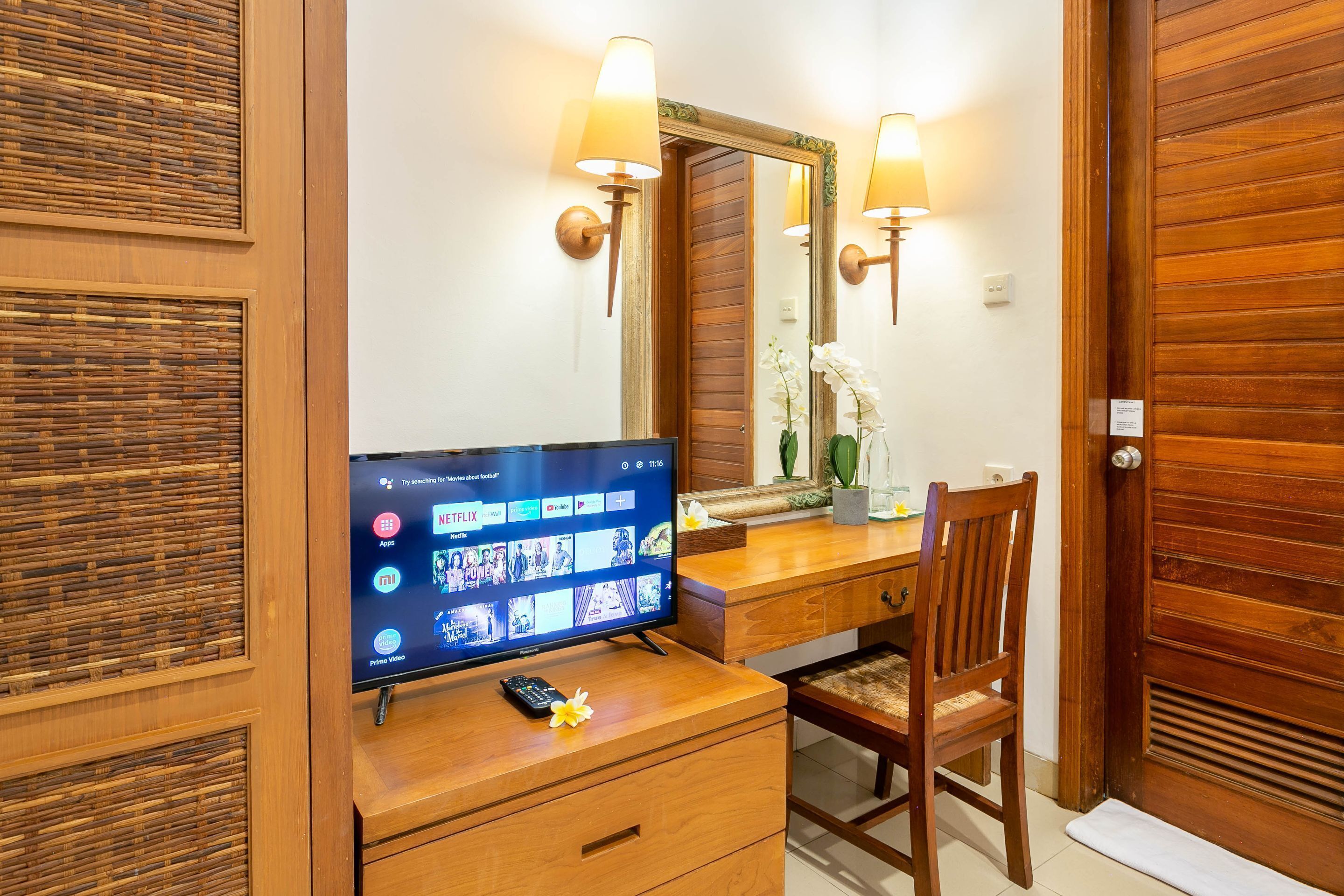 A wooden room with a television on a desk, a chair, and a mirror with wall sconces.