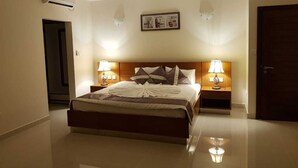 A hotel room with a large bed, two bedside tables with lamps, an air conditioning unit, a ceiling fan, and a framed picture on the wall. - Sahaj Holiday Apartments (Praslin Island)