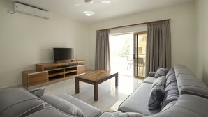 A living room with a grey sofa, a wooden coffee table, a TV on a wooden stand, and a sliding glass door leading to an outdoor area. - Sahaj Holiday Apartments (Praslin Island)