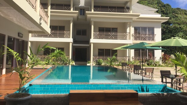 A multi-story hotel with a swimming pool, outdoor seating, and a view of the surrounding greenery. - Sahaj Holiday Apartments (Praslin Island)