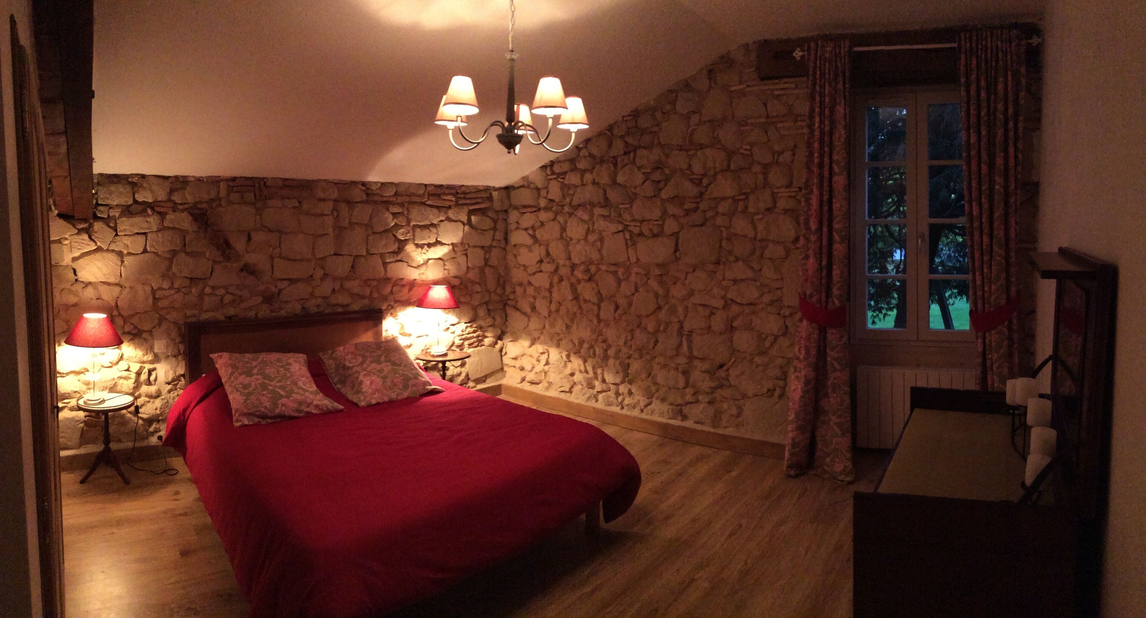 Een slaapkamer met een stenen muur, een bed met rode beddengoed, twee bedlampjes en een raam met gordijnen.
