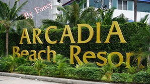 Schild für das Arcadia Beach Resort mit großen, goldenen Lettern auf schwarzem Grund, umgeben von Grünpflanzen.