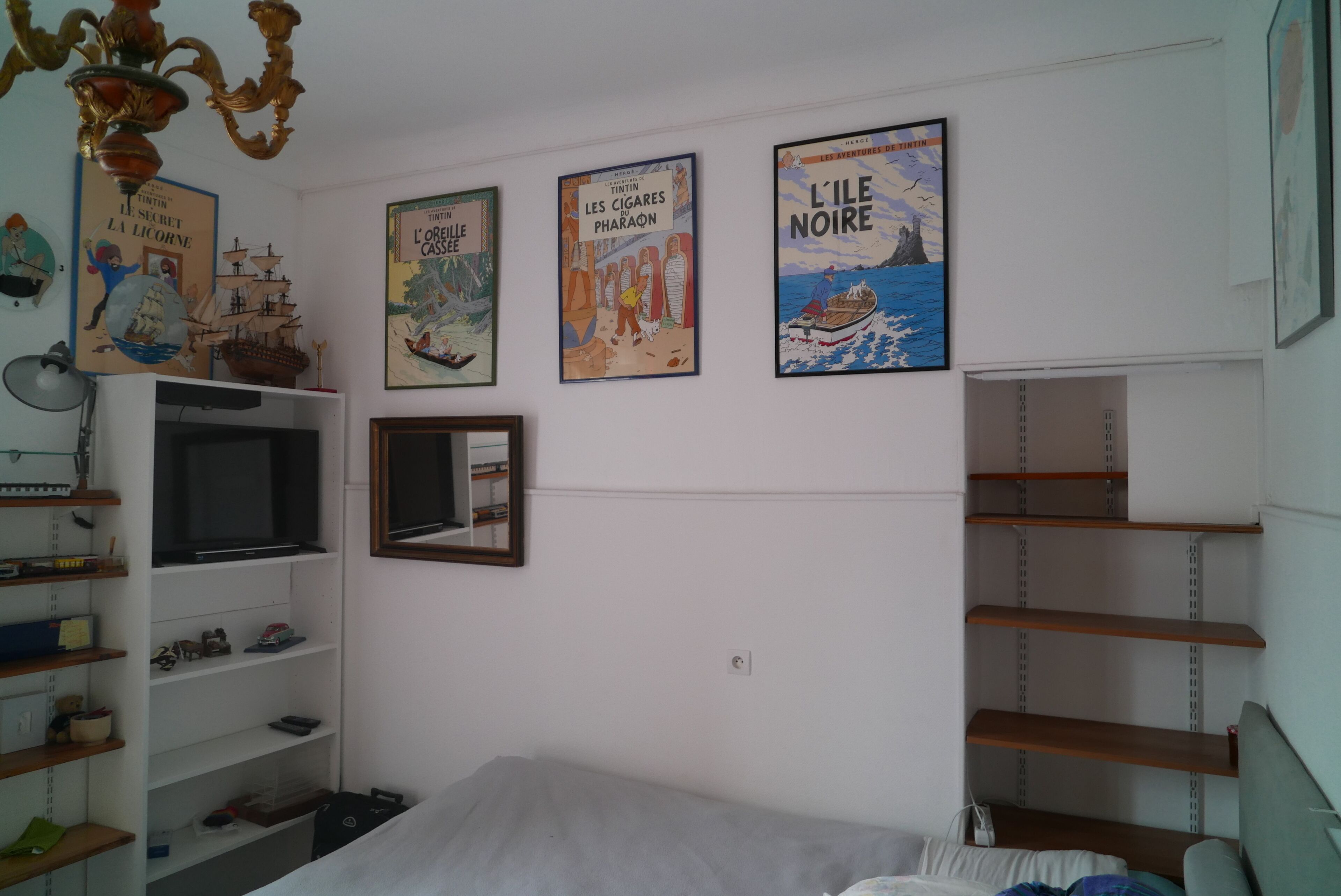 Un dormitorio con cama, estanterías, televisor y tres carteles en la pared.