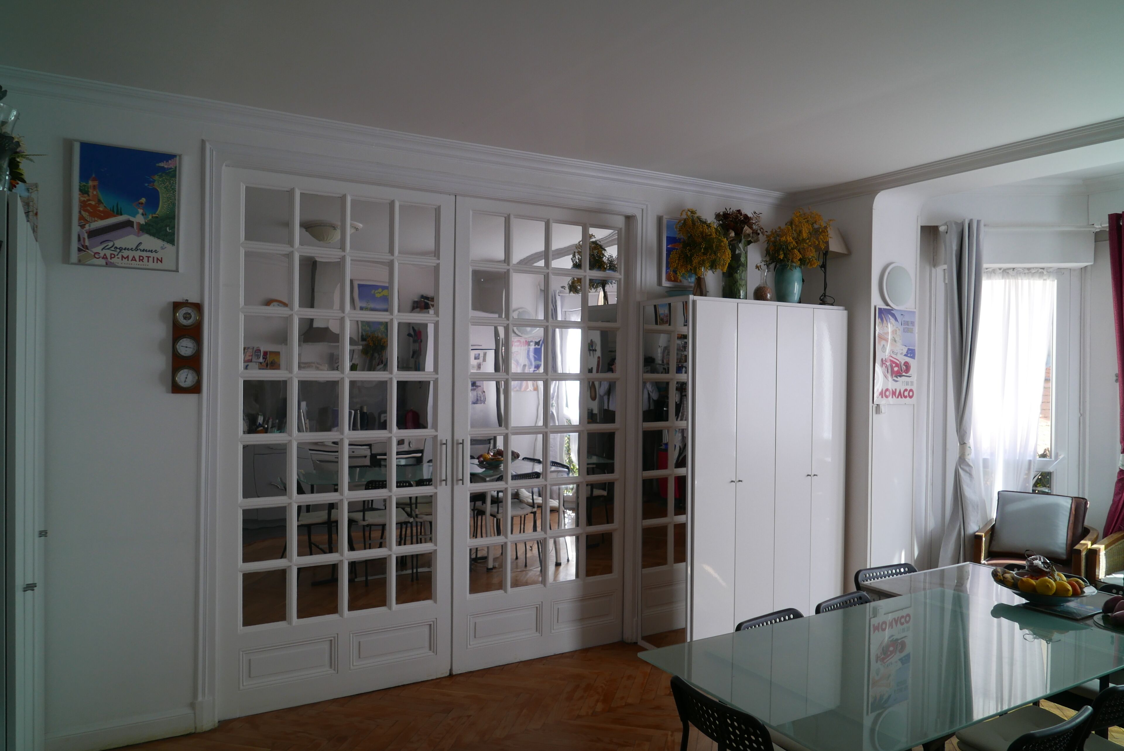 A dining area with a glass table, white cabinets, and a view into another room.