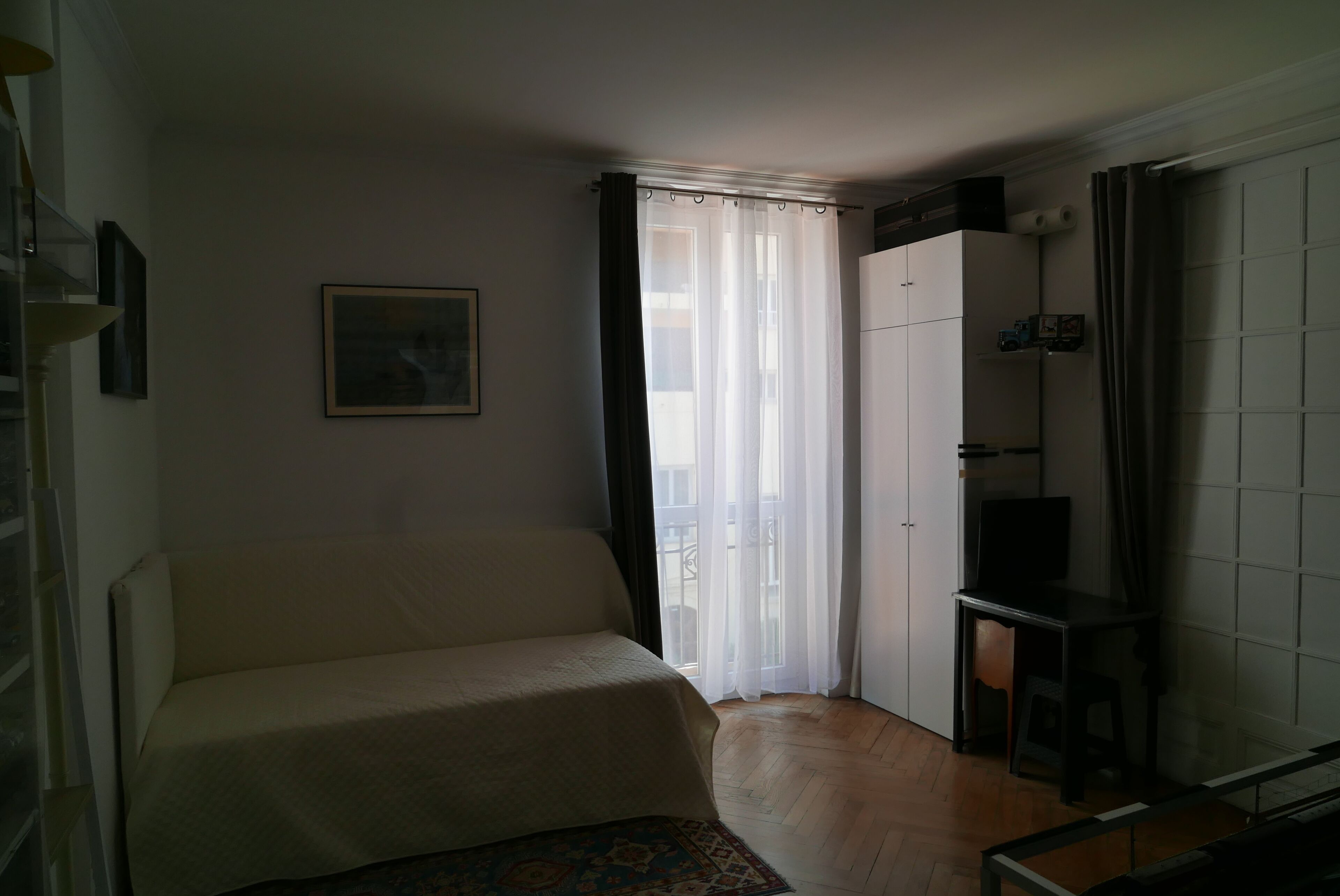Ein kleiner, gut beleuchteter Raum mit einem Bett, einem Fernseher und einem Fenster mit transparenten Vorhängen.