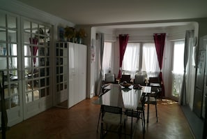 A spacious dining area with a glass table and chairs, white cabinets, and a view of the outdoors through large windows with red curtains.