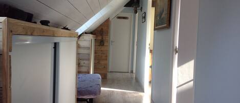 Un pasillo estrecho con un banco de madera, una cama y una puerta que da a otra habitación.