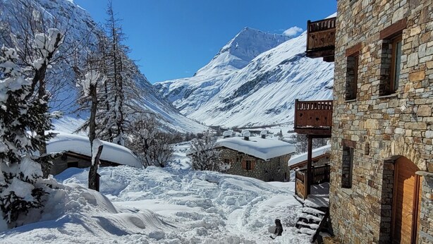 Un village de montagne enneigé, avec des bâtiments en pierre et un ciel bleu azur.