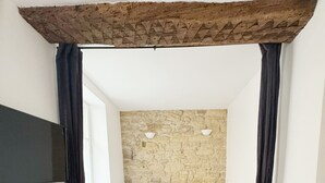A bedroom with a stone wall, a bed with white and yellow pillows, a grey sofa, and a flat-screen TV on the wall. - Suit Du Lion/AC Historic Flat, Marais, 4-5 Guests (Paris)