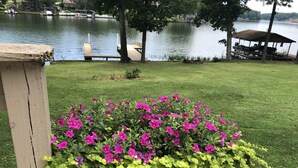 Un jardín con un cantero de madera lleno de flores vibrantes y follaje verde, una terraza de madera y un lago al fondo.