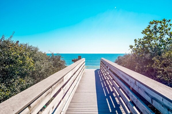 A wooden boardwalk leading to a beach with clear blue water and a bright sky.