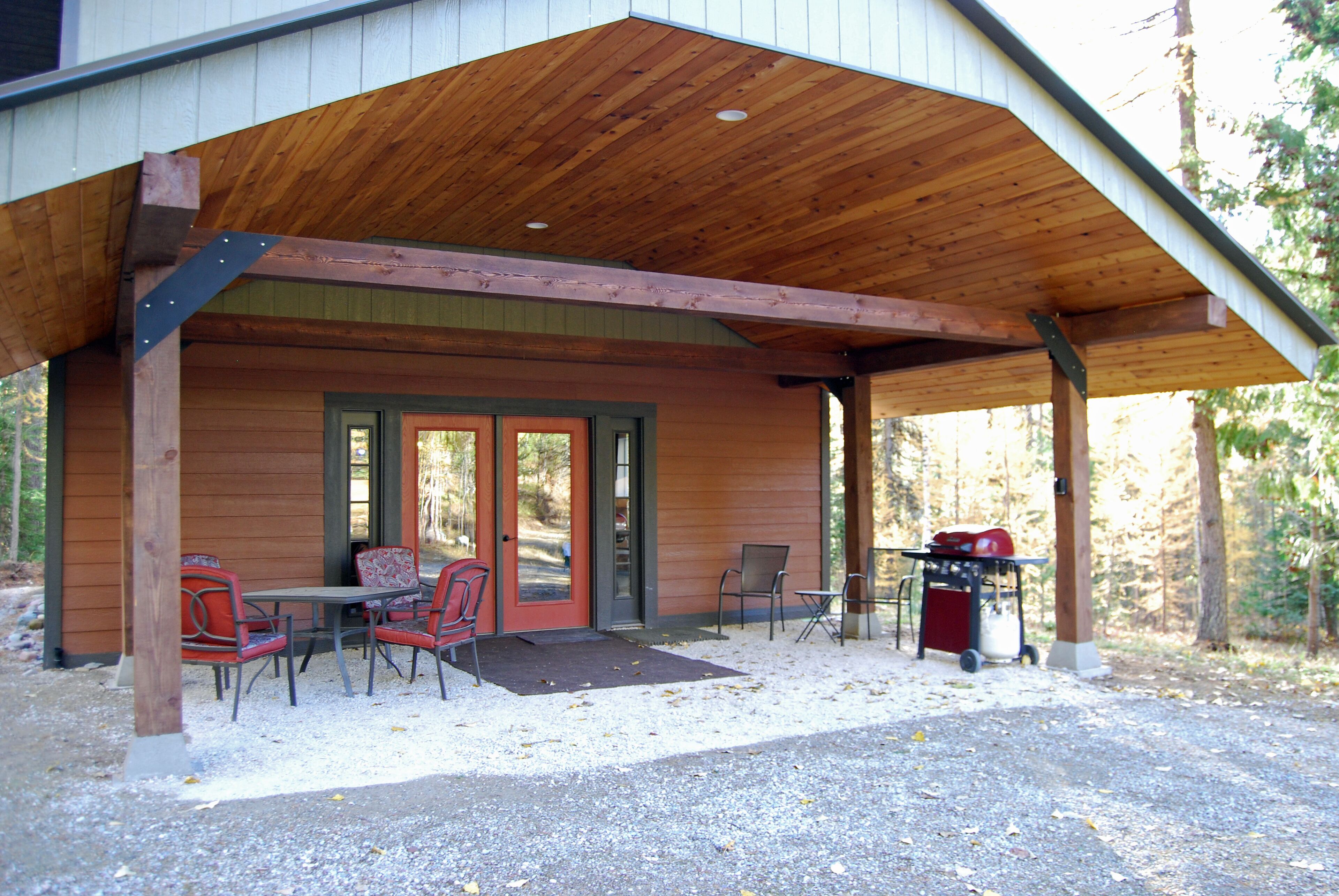 A covered outdoor seating area with tables and chairs, a barbecue grill, and a wooden building with a sloped roof.
