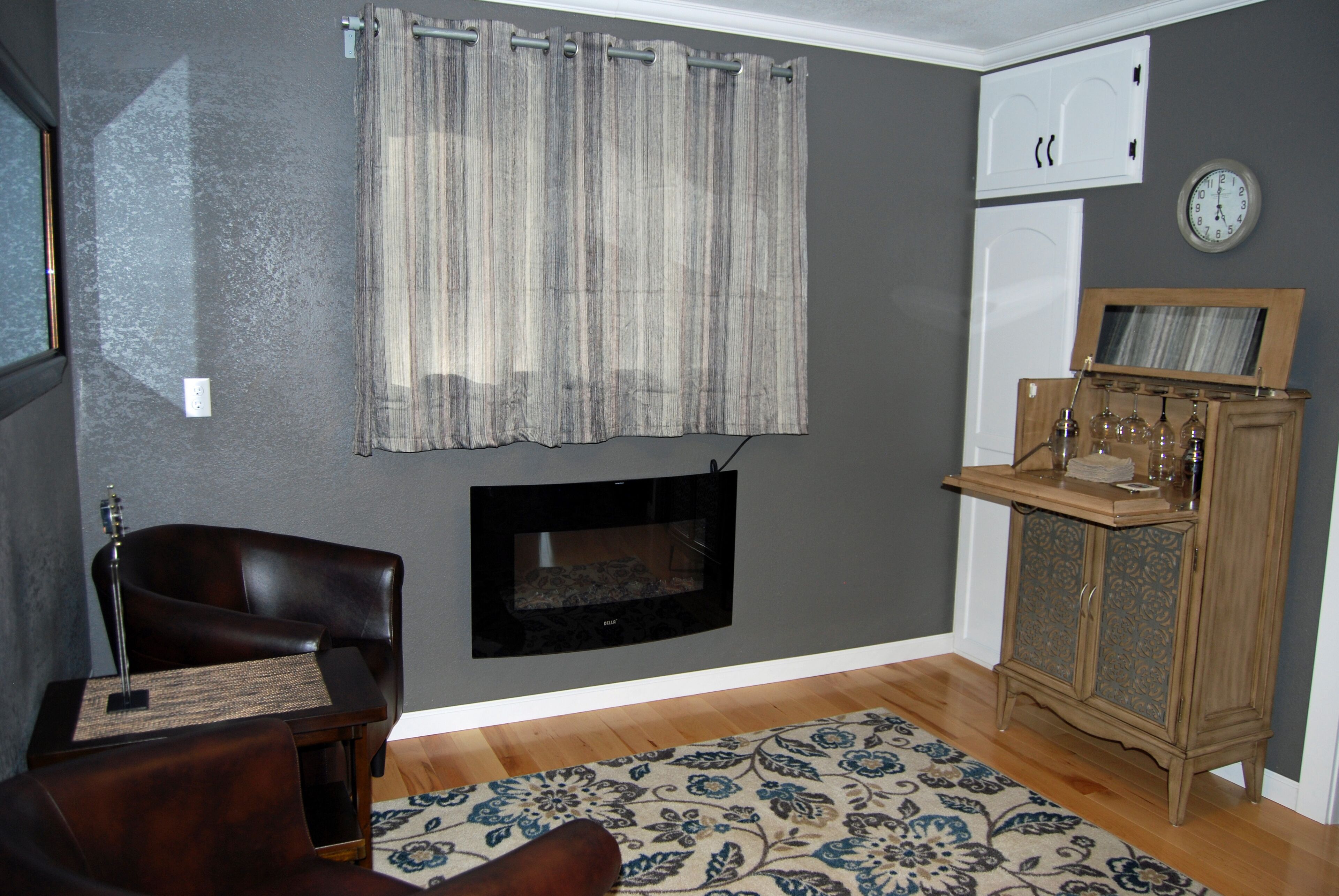 A living room with a fireplace, a dark leather armchair, a patterned rug, and a wooden cabinet with glassware.