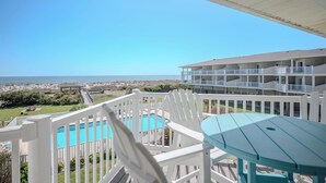 A balcony with a pool, white chairs, and a table overlooking the beach and ocean. - SSV 207: Oceanfront Condo with Pool and Views! (Oak Island)