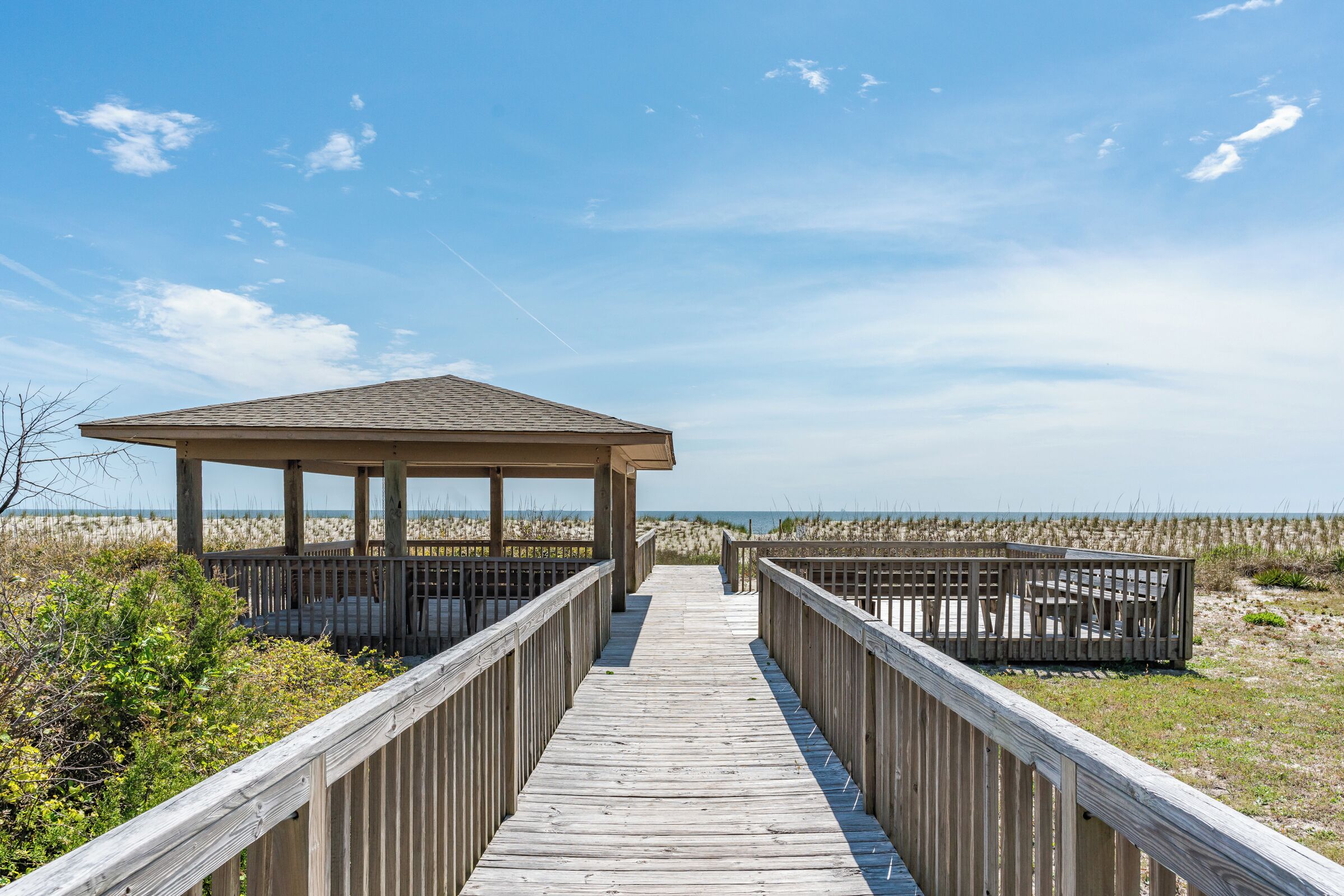 A wooden boardwalk leading to a beach pavilion with a roof, surrounded by sand dunes.
