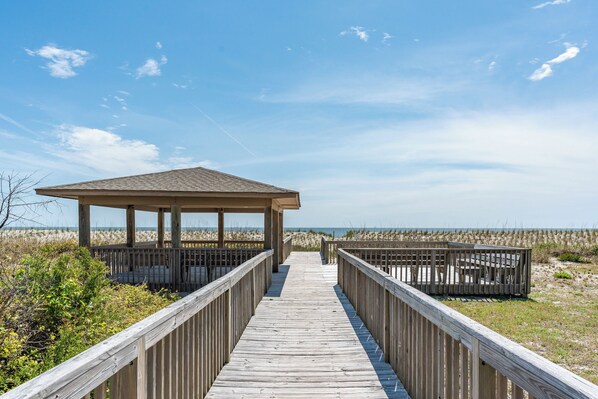 A wooden boardwalk leading to a beach pavilion with a roof, surrounded by sand dunes.