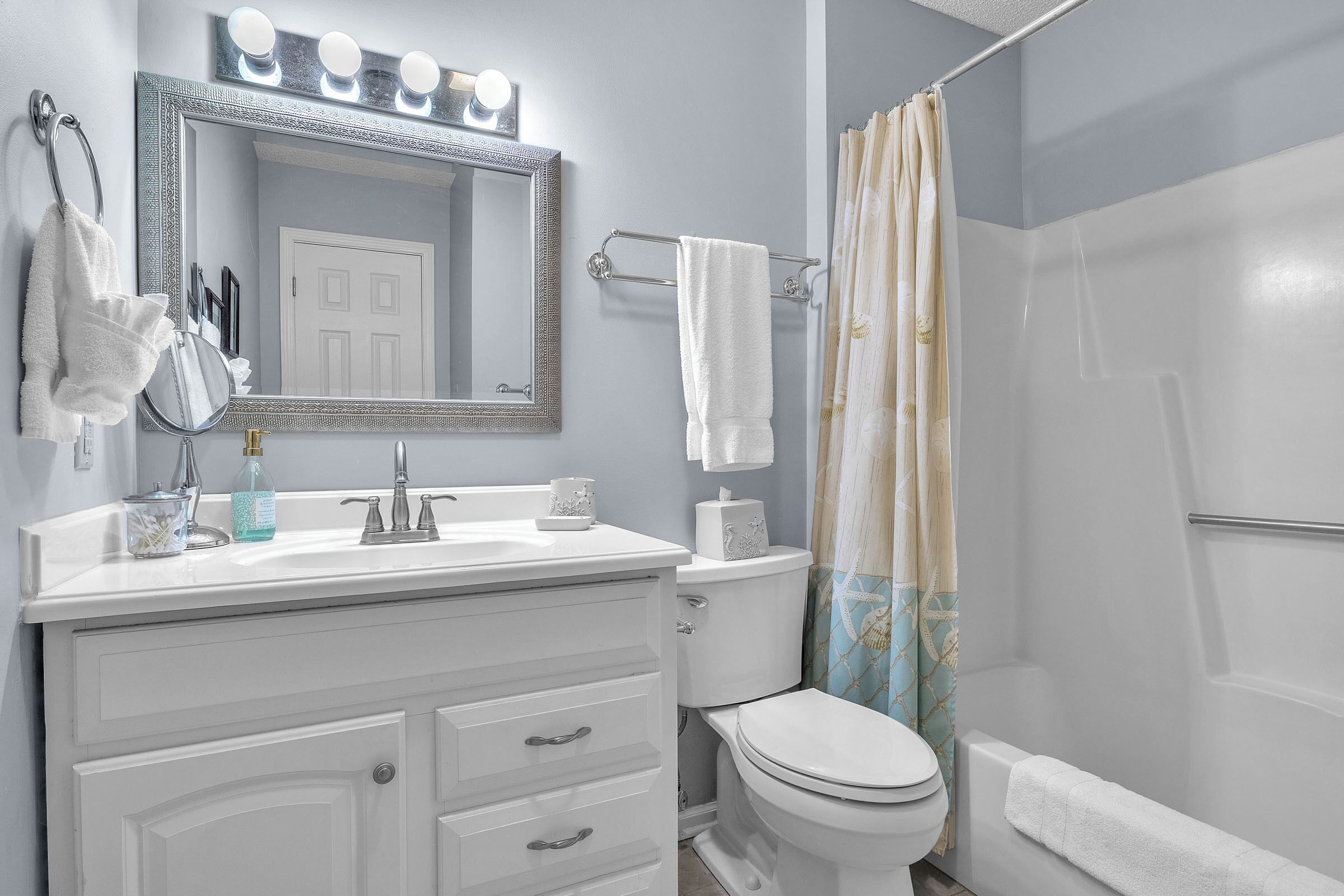 Une salle de bain avec un meuble-lavabo blanc, un grand miroir avec éclairage, des toilettes et une douche avec un rideau.
