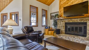 A living room with a stone fireplace, leather sofa, and a television mounted above it. - Large Beautiful Townhome, Private Hot Tub, Near Shuttle Route, In Town, WIFI (Winter Park)