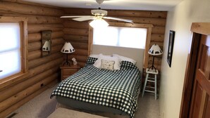 A cozy log cabin bedroom with a bed, two bedside lamps, a ceiling fan, and a window. - Spacious Lake Georgetown Log Home (Anaconda)