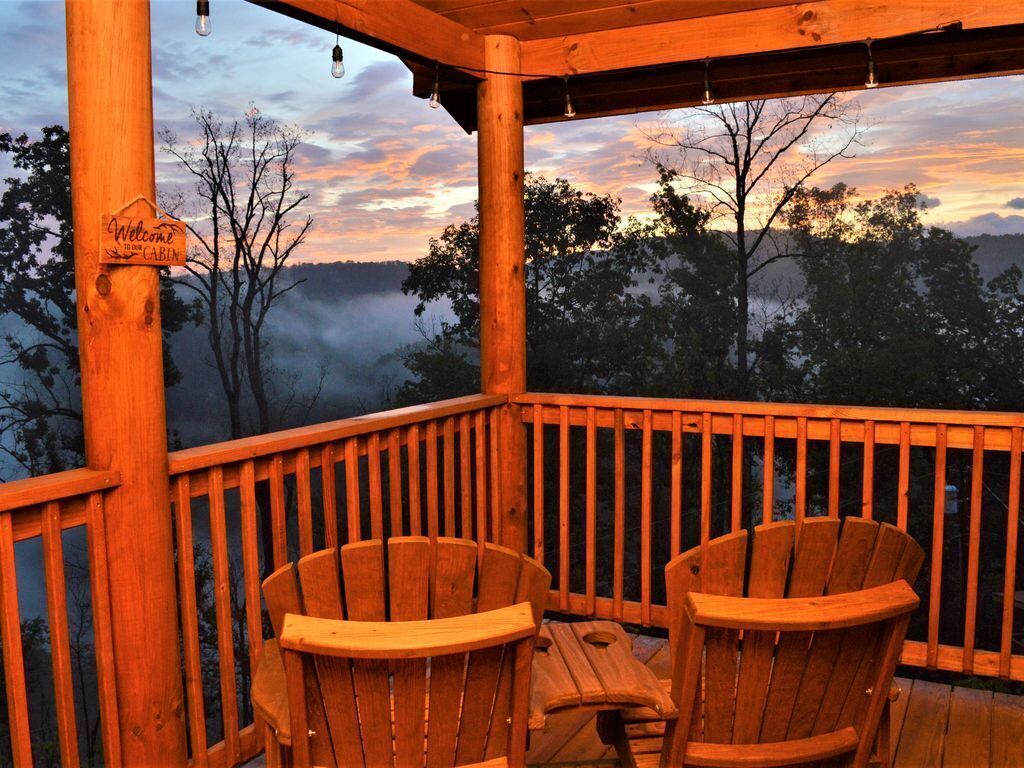 Una terraza de madera con dos sillas Adirondack y una mesa, con vistas a un paisaje boscoso al atardecer.
