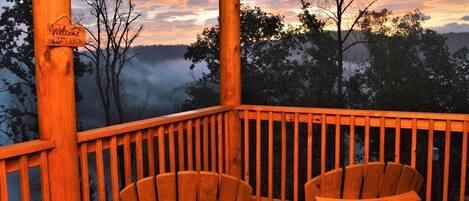 Una terraza de madera con dos sillas Adirondack y una mesa, con vistas a un paisaje boscoso al atardecer.