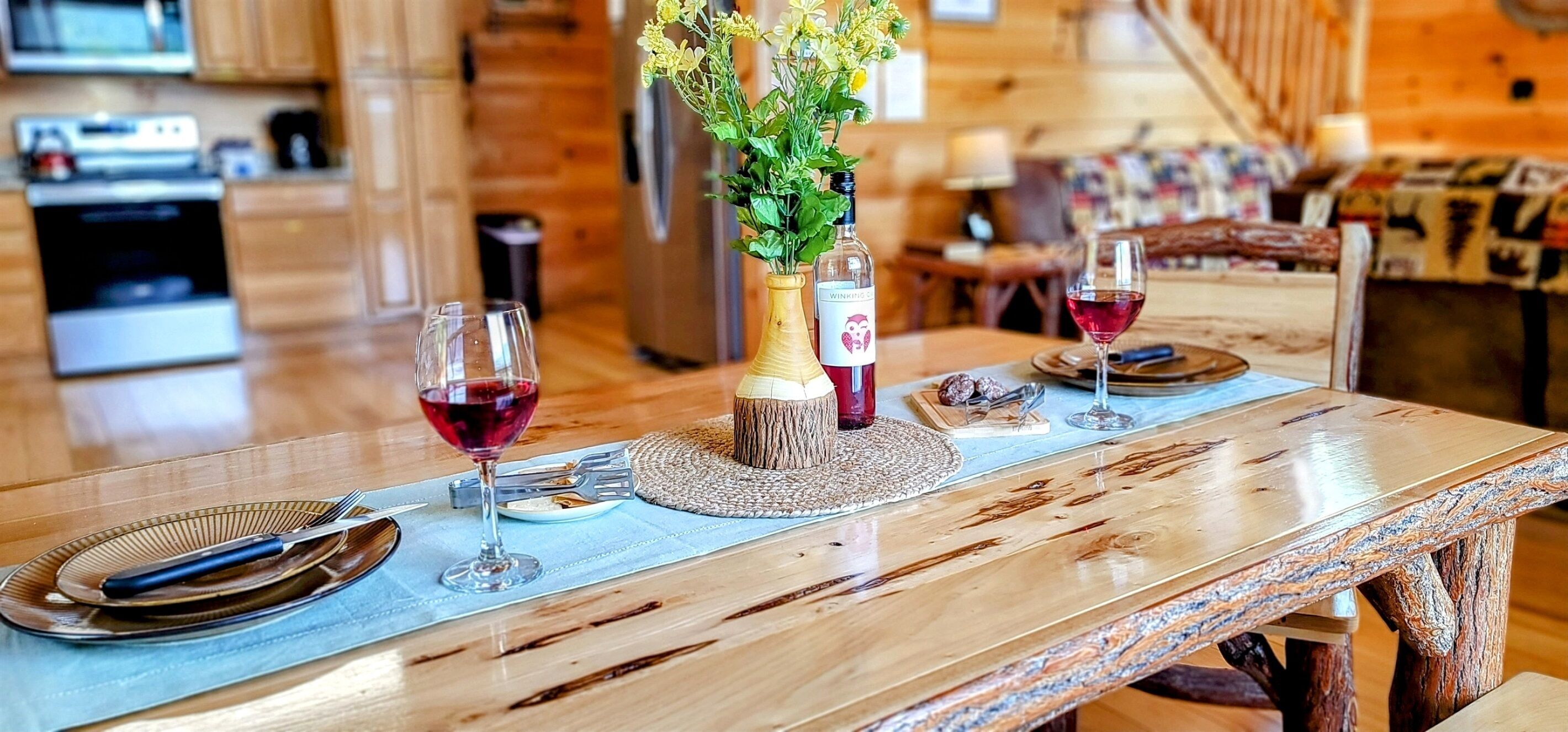 A wooden dining table with a woven placemat, two wine glasses, a bottle of wine, and a vase with flowers.