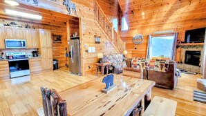 Interior de una cabaña con pisos de madera, una cocina con electrodomésticos modernos, una zona de comedor con mesa y sillas, un sofá y una chimenea.