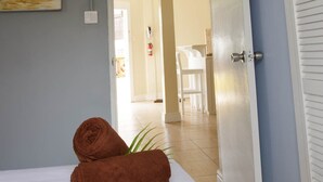 Habitación de hotel con cama, un cuadro de una playa, una toalla y una puerta que da a un comedor.