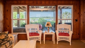 Une pièce aux murs lambrissés, avec deux fauteuils en osier et une table, de grandes fenêtres donnant sur un paysage verdoyant et un bâtiment.