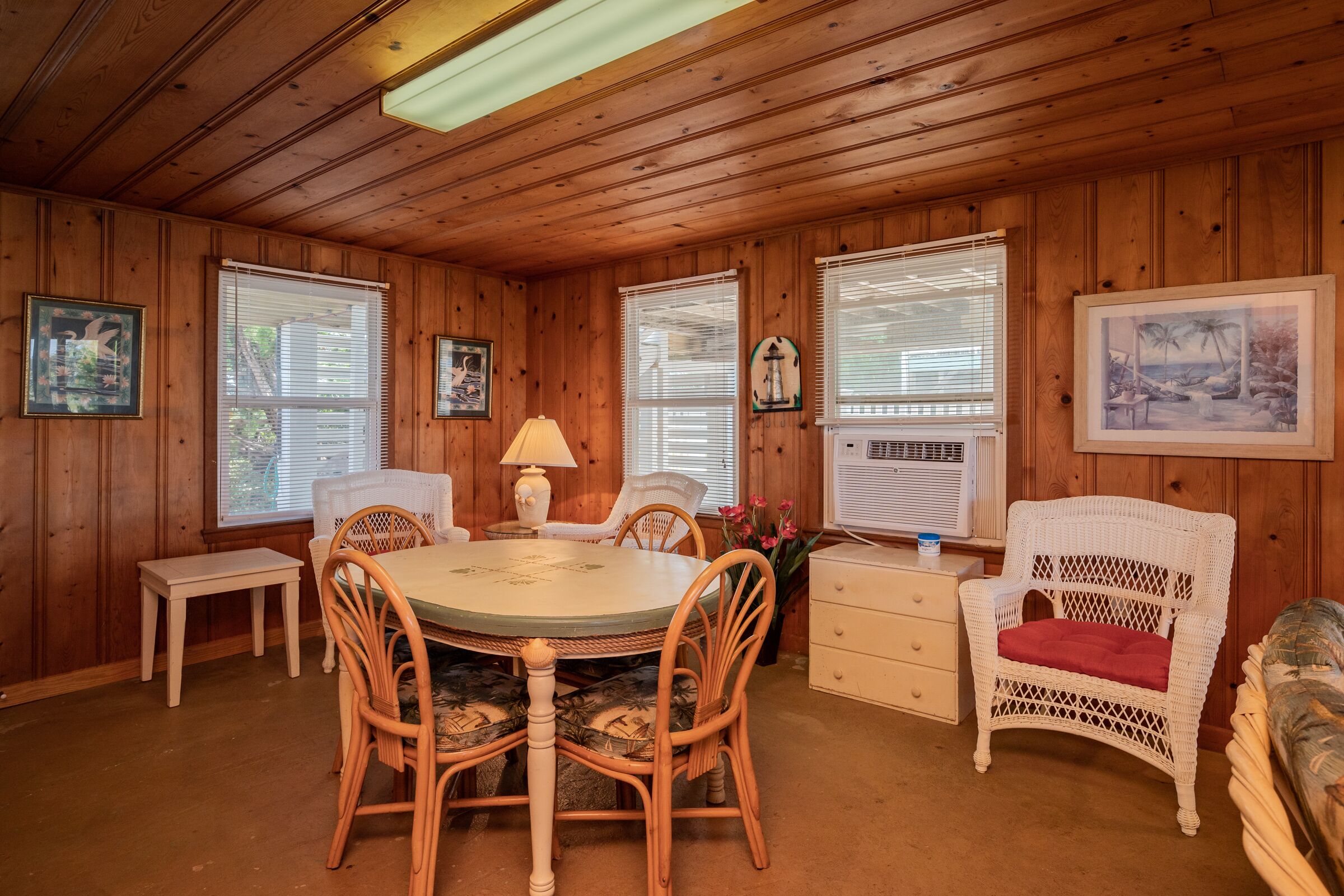 A wooden-paneled room with a round table, chairs, a lamp, a white wicker chair, and framed pictures on the walls.