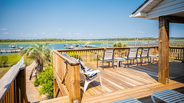 Une terrasse en bois avec des sièges extérieurs, offrant une vue sur une marina avec des bateaux et un ciel bleu dégagé.