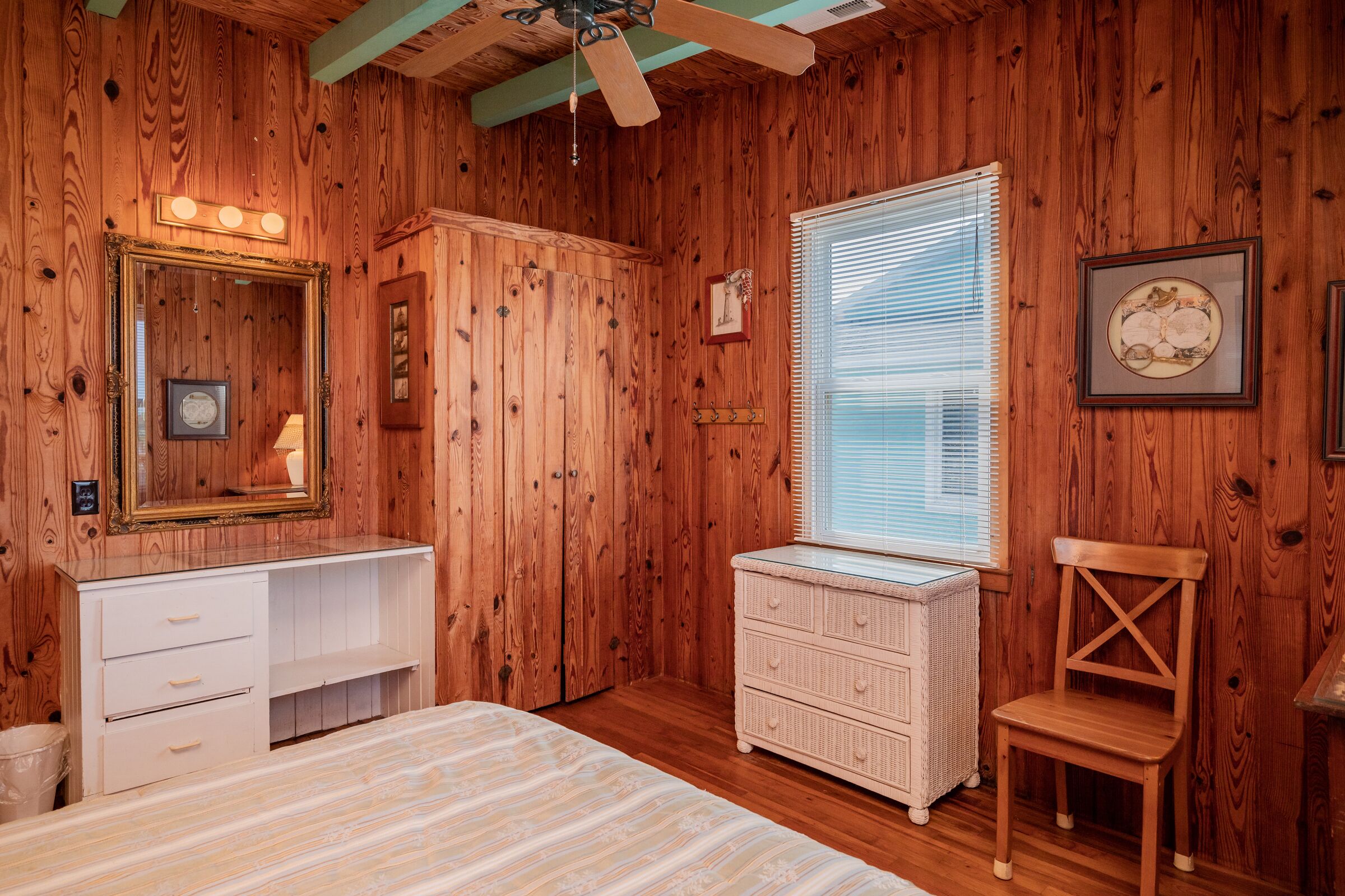 A wooden-paneled room with a bed, a white dresser, a wooden chair, a ceiling fan, and a window with blinds.