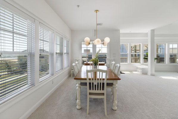 A spacious dining area with a wooden table and white chairs, large windows with blinds, and a modern chandelier. - Sea Lookout - Great last-minute rates with ocean views. June 27-July 4! (Atlantic Beach)