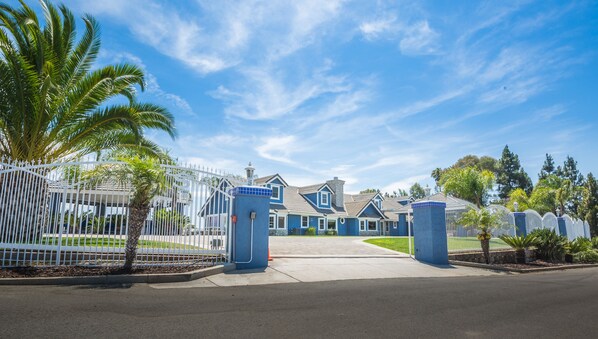 Une entrée fermée par un portail donnant accès à un quartier résidentiel, avec des maisons, des palmiers et un ciel d’un bleu éclatant.
