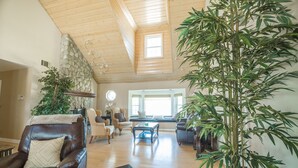 A spacious living room with a high ceiling, wooden beams, and large windows. There are leather chairs, a coffee table, and potted plants. - Hays Family Estate in Temecula (Temecula)