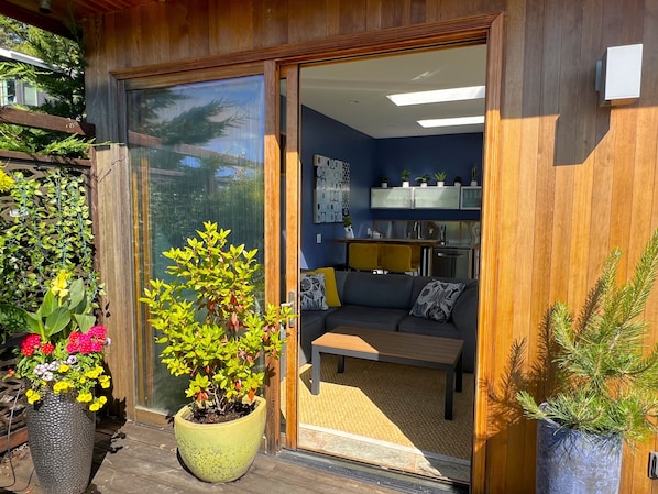 A modern interior with a wooden exterior, a sliding glass door leading to a living area with a sofa and a small table, and a kitchen with cabinets and a countertop.