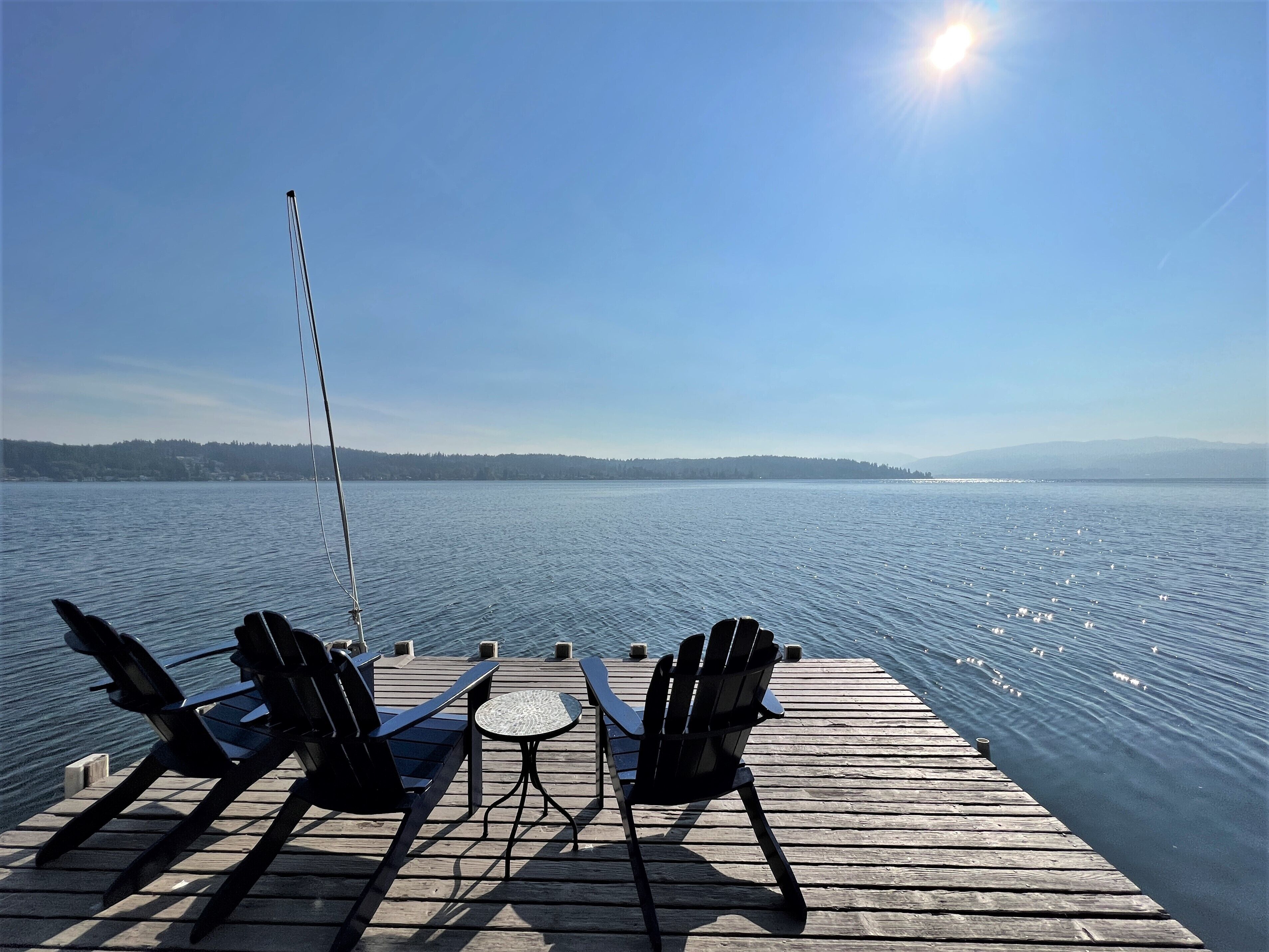 Eine Holzstege mit zwei Stühlen und einem Tisch, der auf einen stillen See mit einer entfernten Uferlinie blickt.
