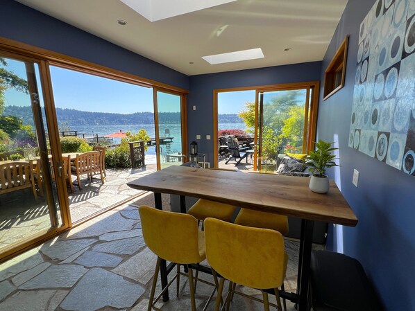 Un comedor con mesa y sillas, ventanas amplias con vista al agua y una planta en maceta en la pared.