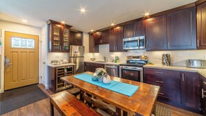 A modern kitchen with dark wooden cabinets, stainless steel appliances, and a wooden dining table with a blue placemat. - Fully Remodeled Condo Near Gondola & Village 276 (Mammoth Lakes)