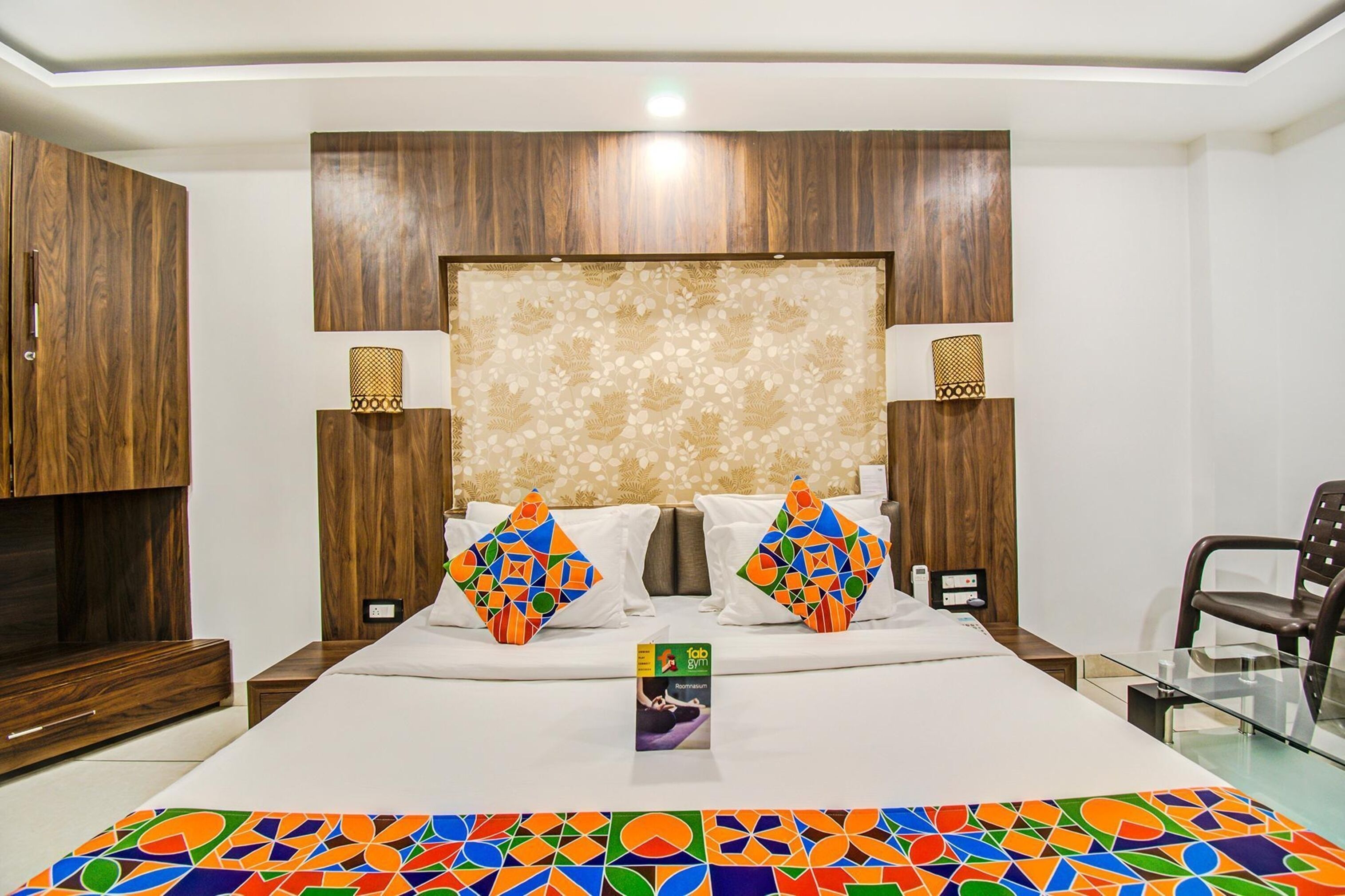 A modern hotel room with a wooden headboard, a large bed with colorful pillows, a glass coffee table, and a TV on a wooden cabinet.