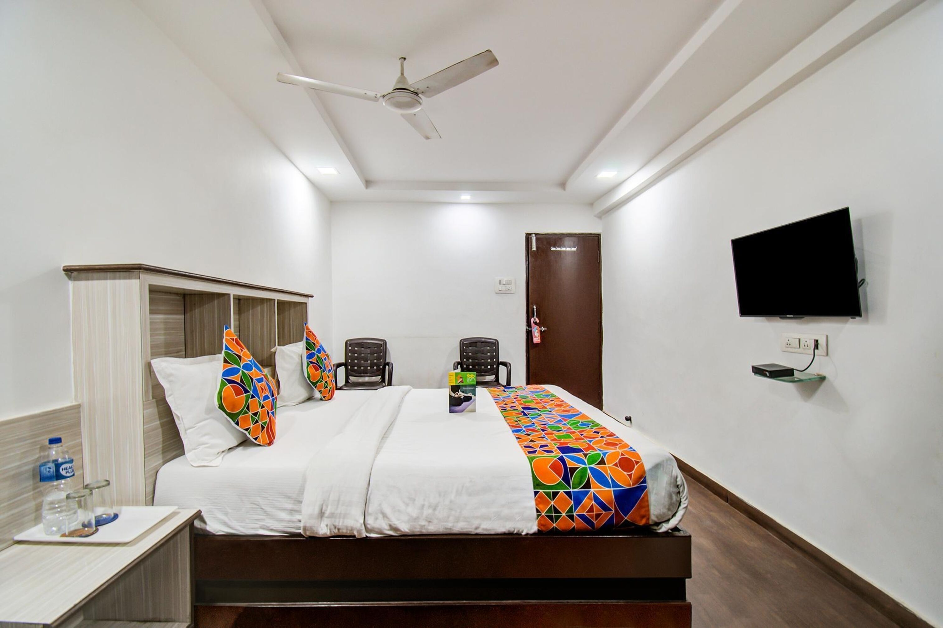 Een hotelkamer met een bed, twee stoelen, een televisie, een plafondventilator en een kledingkast.