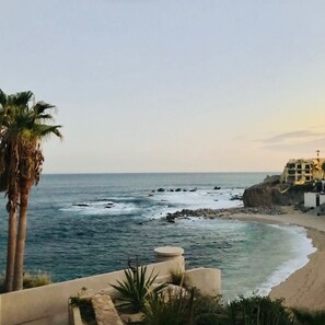 A coastal view with a beach, rocky shoreline, and a building with a balcony overlooking the ocean. - Cozy Mexican Style Cottage  Walking Distance to Beach  Affordable Cabo Retreat for Long and Short Stays (Cabo San Lucas)