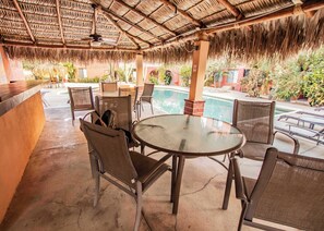 An outdoor seating area with a round glass table and metal chairs, a pool in the background, and a thatched roof overhead. - Cozy Mexican Style Cottage  Walking Distance to Beach  Affordable Cabo Retreat for Long and Short Stays (Cabo San Lucas)