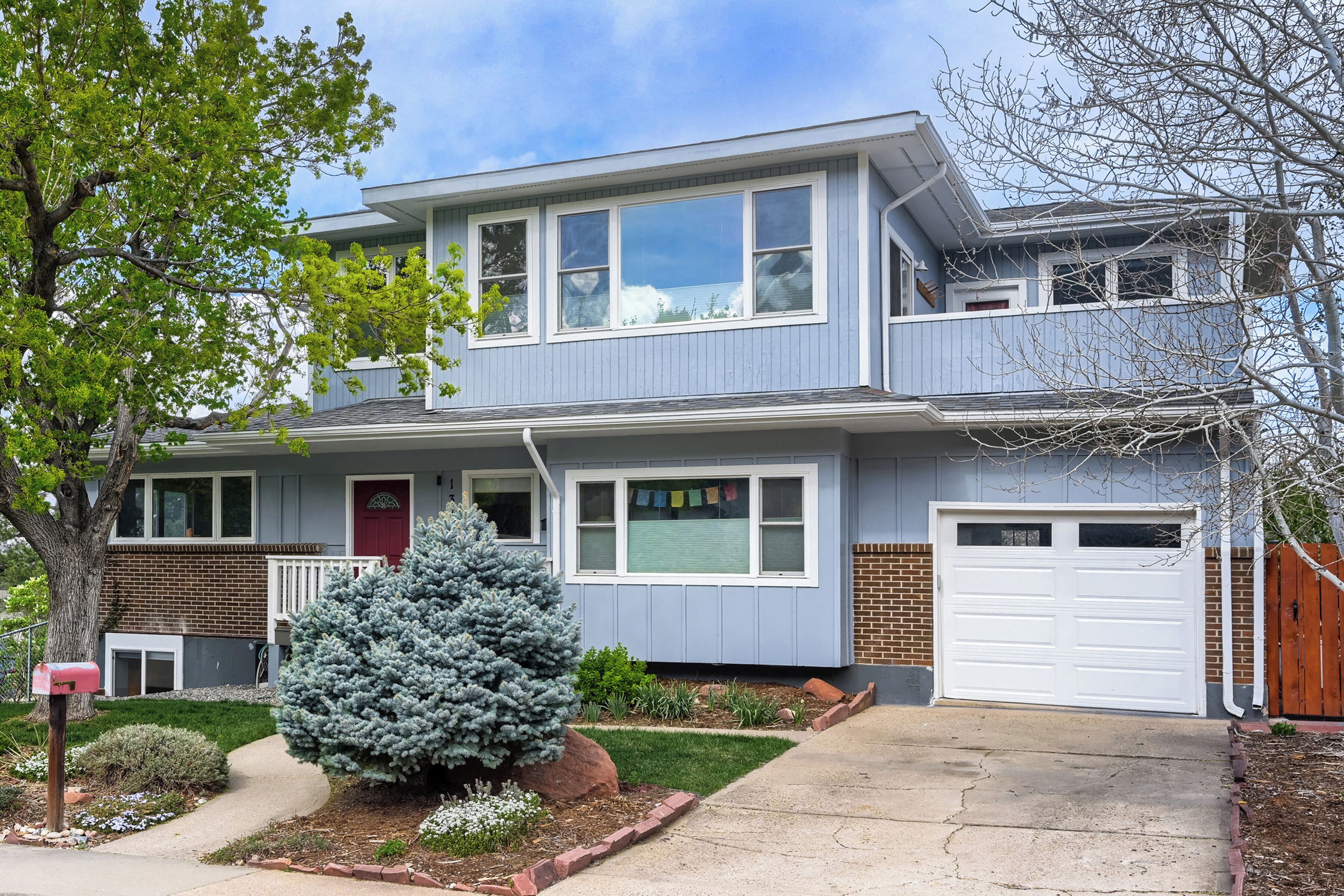 A two-story house with a front garage, a red door, and a small front yard with a coniferous tree and shrubs.